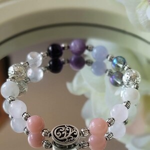 May include: A beaded bracelet featuring a variety of colorful gemstones. The bracelet includes pink, white, purple, and blue beads, interspersed with silver-colored metal accents. The clasp is also silver-toned with an ornate design.