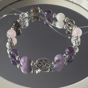 May include: A beaded bracelet featuring a mix of round polished gemstones in shades of purple, pink, clear, and smoky gray, interspersed with small silver-toned metal spacers and a central oval silver-toned charm with an intricate filigree design.