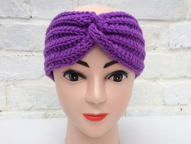 May include: Purple crocheted headband with a knotted design. The headband is worn on a mannequin head.