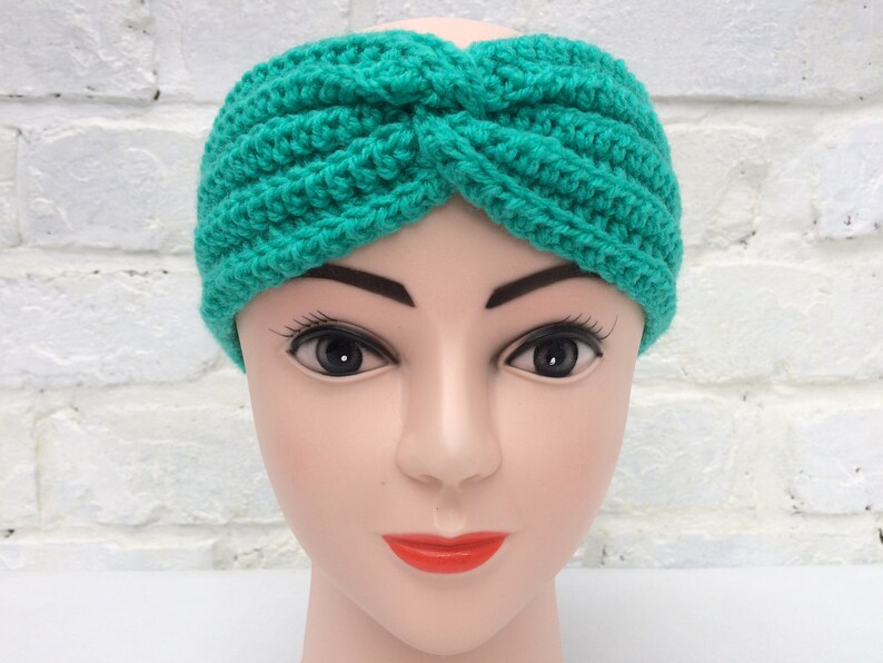 May include: A teal blue crocheted headband with a twist design. The headband is on a mannequin head.