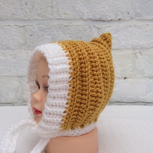 May include: A white and mustard yellow crocheted baby bonnet with a pointed top and ties. The bonnet is on a white mannequin head.
