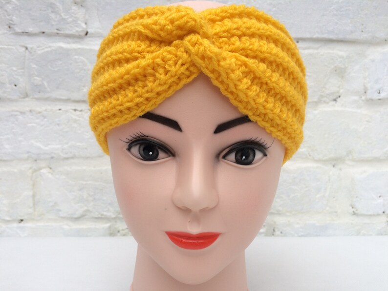 May include: A yellow crocheted headband with a twisted knot design. The headband is worn on a mannequin head.