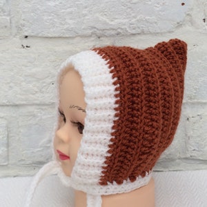 Puede incluir: Un gorro de bebé de ganchillo blanco y marrón con un lazo. El gorro tiene una parte superior puntiaguda y un borde blanco.
