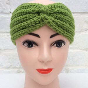 May include: A green crocheted headband with a knotted design. The headband is worn on a mannequin head.