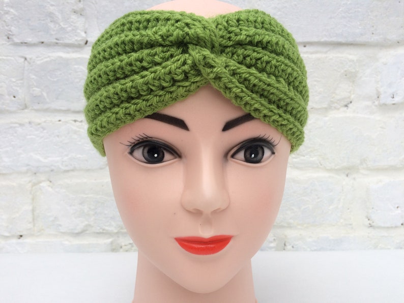 May include: A green crocheted headband with a knotted design. The headband is worn on a mannequin head.