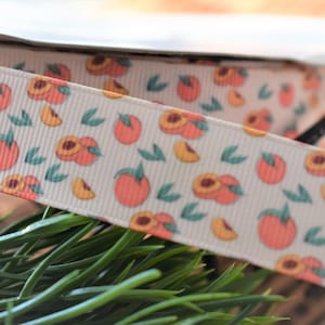 May include: White ribbon with a peach print. The ribbon has a repeating pattern of peaches and leaves. The peaches are a light orange color and the leaves are a light green color.