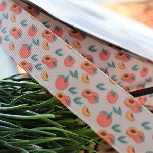 May include: A white ribbon with a peach pattern. The ribbon has a repeating pattern of peaches and leaves. The peaches are pink and orange and the leaves are green.