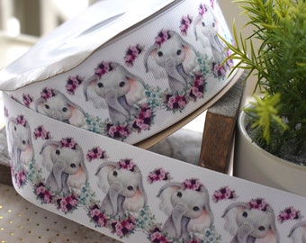 May include: White ribbon spool featuring a repeating pattern of watercolor-style elephants wearing floral crowns. The elephants are gray with pink accents, surrounded by pink and green floral elements. The ribbon is ideal for crafting and gift wrapping.