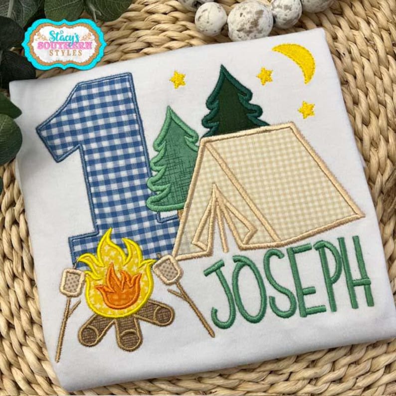 K&ouml;nnte beinhalten: Wei&szlig;es T-Shirt mit Camping-Motiv. Es zeigt eine blau-karierte Zahl 1, ein Lagerfeuer mit Marshmallows, ein Zelt, B&auml;ume, Sterne und einen gelben Mond. Der Name "JOSEPH" ist gr&uuml;n gestickt.