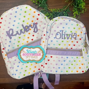 May include: Two white backpacks with colorful heart patterns. One backpack has the name "Ruby" embroidered in purple, and the other has "Olivia." Both have purple zippers and straps. A tag with the text "Stacy's Southern Styles" is attached.