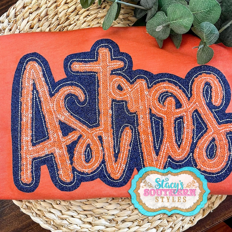 May include: Orange and navy blue sequined shirt with the word "Ashlyn" in a cursive font. The shirt is on a woven straw surface. The image also includes a small tag with the text "Stacy's Southern Styles".