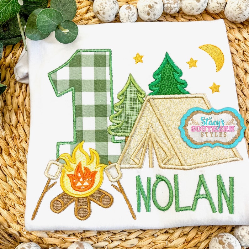 K&ouml;nnte beinhalten: Wei&szlig;es T-Shirt mit Camping-Motiv. Es zeigt die gr&uuml;ne karierte Zahl 1, ein Lagerfeuer mit Marshmallows, ein Zelt, B&auml;ume, Sterne und einen Mond. Der Name "NOLAN" ist gr&uuml;n gestickt. Mit dem Text "Stacy's SOUTHERN STYLES".
