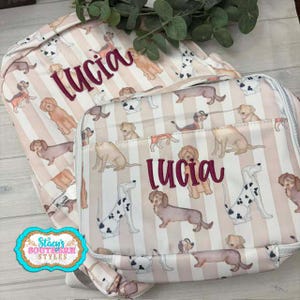 May include: A backpack and a rectangular lunch bag featuring a beige and white vertical striped pattern with various dog breeds printed throughout. Both items are personalized with the name Lucia embroidered in a dark red script font.