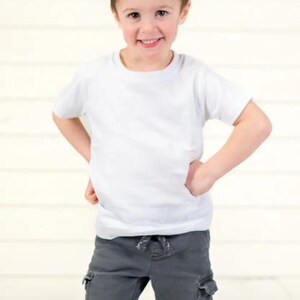 Pu&ograve; includere: Un bambino piccolo con una maglietta bianca e pantaloni cargo grigi, sorridente con le mani sui fianchi. L'outfit &egrave; casual e adatto all'uso quotidiano. Lo sfondo &egrave; di colore chiaro e neutro.