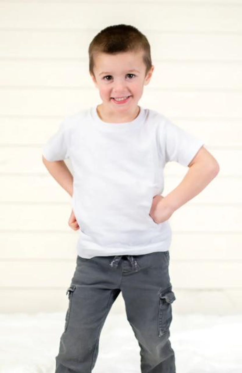 Pu&ograve; includere: Un bambino piccolo con una maglietta bianca e pantaloni cargo grigi, sorridente con le mani sui fianchi. L'outfit &egrave; casual e adatto all'uso quotidiano. Lo sfondo &egrave; di colore chiaro e neutro.