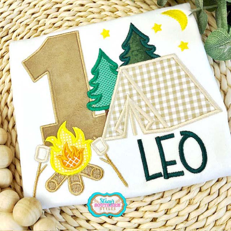K&ouml;nnte beinhalten: Wei&szlig;es T-Shirt mit Camping-Motiv. Mit einer gro&szlig;en Zahl 1, einem Zelt, B&auml;umen, einem Lagerfeuer mit Marshmallows und dem Namen "LEO" in Gr&uuml;n. Enth&auml;lt einen gelben Mond und Sterne. Das Logo "Stacy's Southern Styles" ist sichtbar.