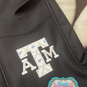 May include: Black bag with a white embroidered "A T M" design. The letters are filled with a floral pattern of white and blue flowers. A sticker with the text "Stacy's Southern Styles" is on the bag.