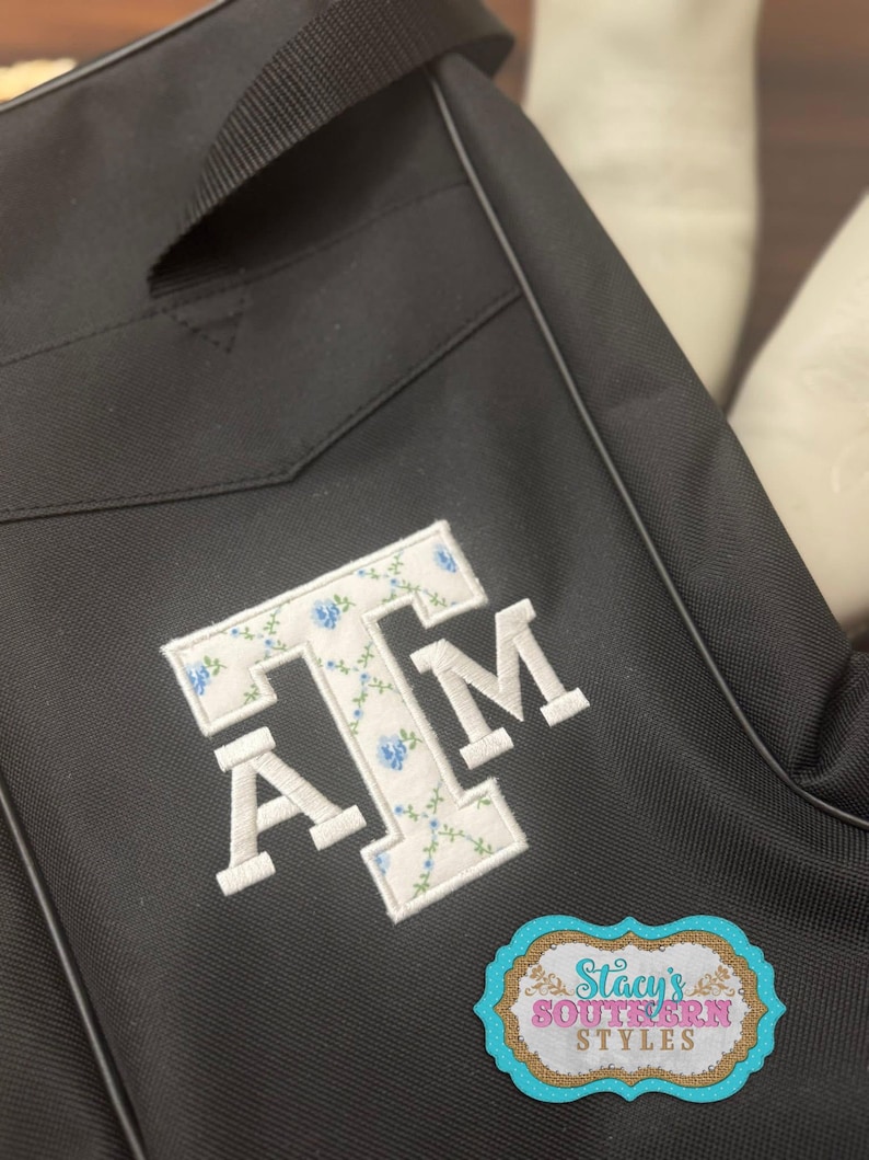 May include: Black bag with a white embroidered "A T M" design. The letters are filled with a floral pattern of white and blue flowers. A sticker with the text "Stacy's Southern Styles" is on the bag.