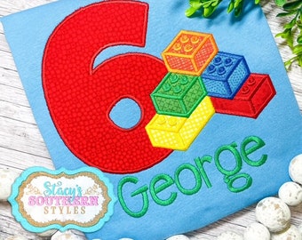 May include: A blue t-shirt with a red number six and a stack of colorful toy building blocks. The name "George" is embroidered in green below the blocks. The shirt has a small logo for "Stacy's Southern Styles" in the bottom left corner.