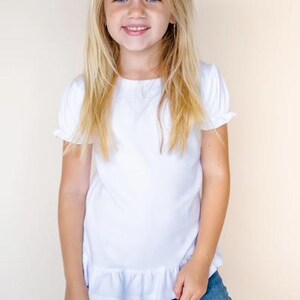 Pu&ograve; includere: Una ragazza con lunghi capelli biondi indossa un top bianco a maniche corte con orlo a balze e jeans blu. Ha un fiocco azzurro chiaro tra i capelli.