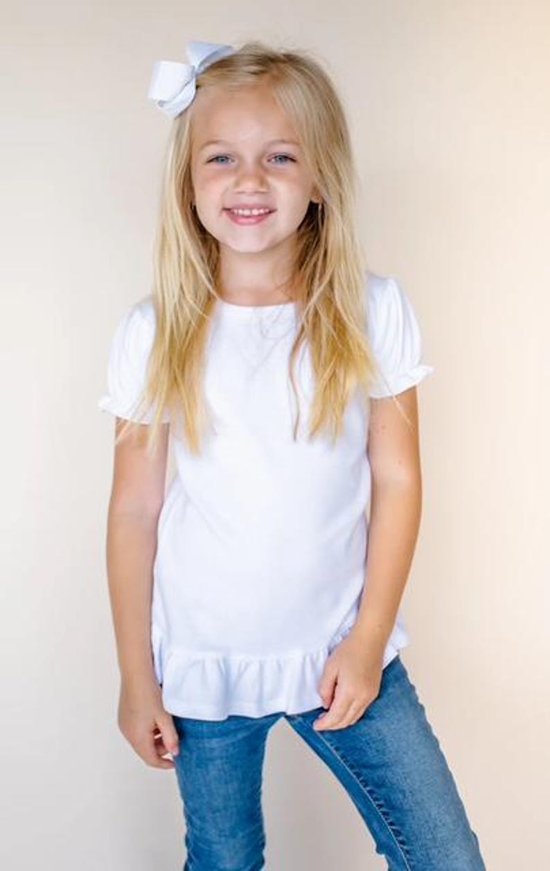 Pu&ograve; includere: Una ragazza con lunghi capelli biondi indossa un top bianco a maniche corte con orlo a balze e jeans blu. Ha un fiocco azzurro chiaro tra i capelli.