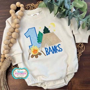 May include: Cream-colored baby romper with a camping theme. Features a blue gingham number 1, a tent, trees, a campfire, and the name "BANKS" in blue. The romper has long sleeves and a round neckline. The text "Stacy's Southern Styles" is also visible.