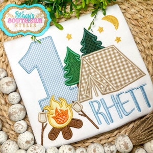 K&ouml;nnte beinhalten: Wei&szlig;es T-Shirt mit Camping-Motiv. Es zeigt eine blau-wei&szlig; karierte Zahl 1, ein Zelt, B&auml;ume, ein Lagerfeuer mit Marshmallows und den Namen "Rhett" in Blau. Enth&auml;lt einen goldenen Mond und Sterne. Das Shirt hat den Text "Stacy's Southern Styles".