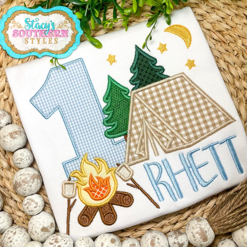 K&ouml;nnte beinhalten: Wei&szlig;es T-Shirt mit Camping-Motiv. Es zeigt eine blau-wei&szlig; karierte Zahl 1, ein Zelt, B&auml;ume, ein Lagerfeuer mit Marshmallows und den Namen "Rhett" in Blau. Enth&auml;lt einen goldenen Mond und Sterne. Das Shirt hat den Text "Stacy's Southern Styles".