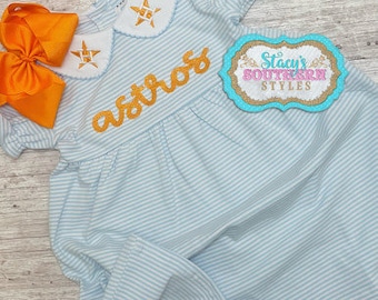 May include: A light blue and white striped dress with a white Peter Pan collar and gold embroidery that says "astros". The dress has a star design on the collar and a bow on the front.