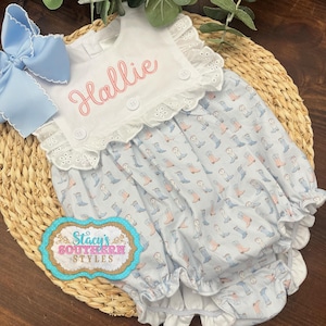 May include: A baby romper with a blue and white boot print, a white eyelet collar, and the name "Hallie" embroidered in pink. A large blue bow is attached. The romper is displayed on a woven mat.