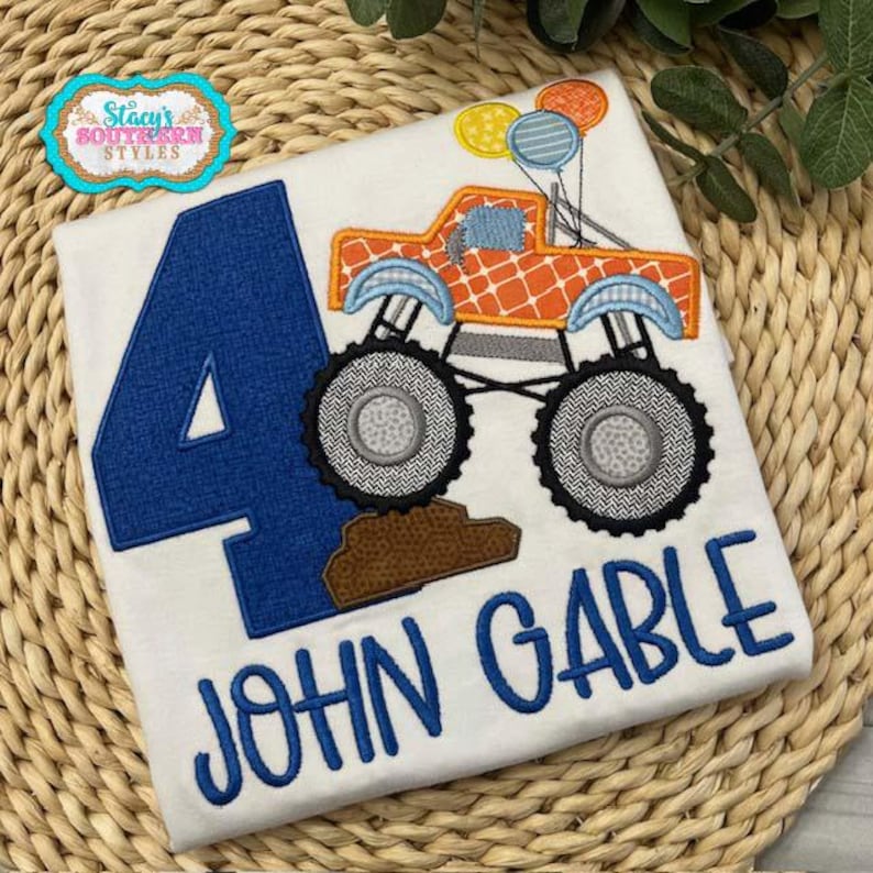 Pu&ograve; includere: T-shirt bianco con un numero 4 blu e il nome "JOHN GABLE" in blu. Un monster truck arancione con dettagli blu e palloncini &egrave; ricamato sulla maglietta. La maglietta &egrave; decorata con terra marrone.