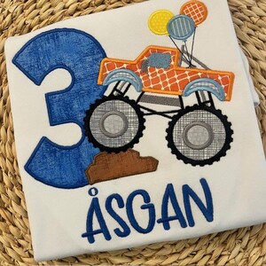 Pu&ograve; includere: T-shirt bianco con un numero 3 blu e il nome "&Aring;SGAN" in blu. &Egrave; presente anche un monster truck arancione con palloncini. Il camion ha grandi ruote nere ed &egrave; decorato con vari motivi.
