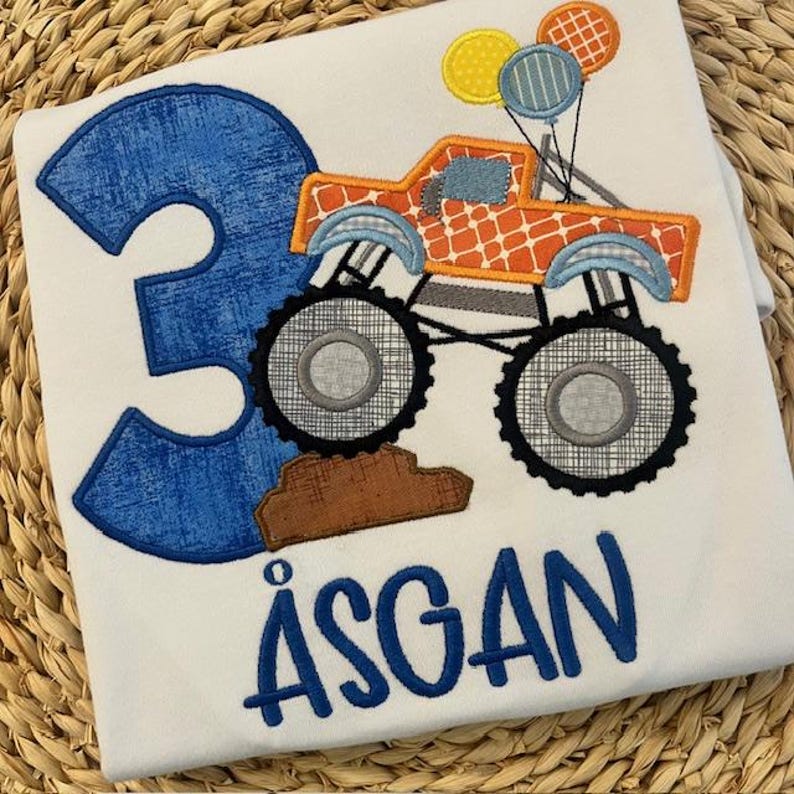 Pu&ograve; includere: T-shirt bianco con un numero 3 blu e il nome "&Aring;SGAN" in blu. &Egrave; presente anche un monster truck arancione con palloncini. Il camion ha grandi ruote nere ed &egrave; decorato con vari motivi.