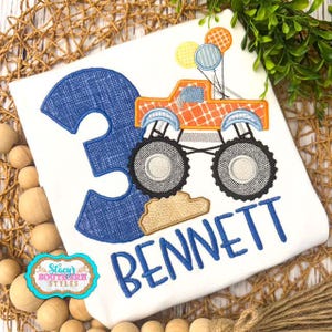 Pu&ograve; includere: T-shirt bianco con il numero 3 blu e il nome "BENNETT" in blu. Un monster truck arancione con palloncini &egrave; sulla maglietta. La maglietta ha anche il testo "Stacy's Southern Styles" in un emblema turchese e oro.