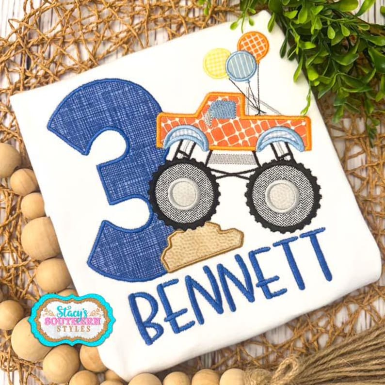 Pu&ograve; includere: T-shirt bianco con il numero 3 blu e il nome "BENNETT" in blu. Un monster truck arancione con palloncini &egrave; sulla maglietta. La maglietta ha anche il testo "Stacy's Southern Styles" in un emblema turchese e oro.