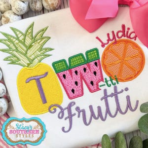 May include: A white t-shirt with the text "Two Frutti" embroidered in purple. The "T" in "Two" is a yellow pineapple, the "W" is a pink watermelon, and the "i" is an orange slice. The name "Lydia" is embroidered in purple above the fruit.