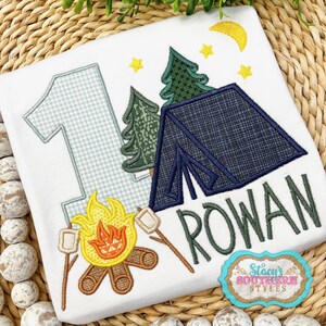 K&ouml;nnte beinhalten: Wei&szlig;es T-Shirt mit Camping-Motiv. Es zeigt ein blaues Zelt, gr&uuml;ne B&auml;ume, ein Lagerfeuer mit Marshmallows und den Namen "Rowan" in Gr&uuml;n. Enth&auml;lt die Zahl "1" und einen Mond.