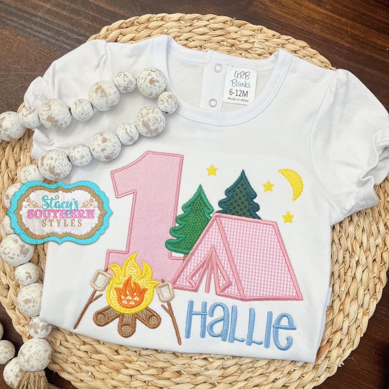 K&ouml;nnte beinhalten: Wei&szlig;es Kurzarm-T-Shirt mit Camping-Motiv. Es zeigt eine rosa Zahl 1, ein Zelt, B&auml;ume, ein Lagerfeuer mit Marshmallows und den Namen "Hallie" in Blau. Enth&auml;lt den Text "CRB Blanks 6-12M".