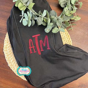 May include: Black bag with a red embroidered "ATM" monogram. The bag has a zipper closure and a light-colored trim. The bag is decorated with a sprig of green leaves and a small, colorful sticker that says "Stacy's Southern Styles".