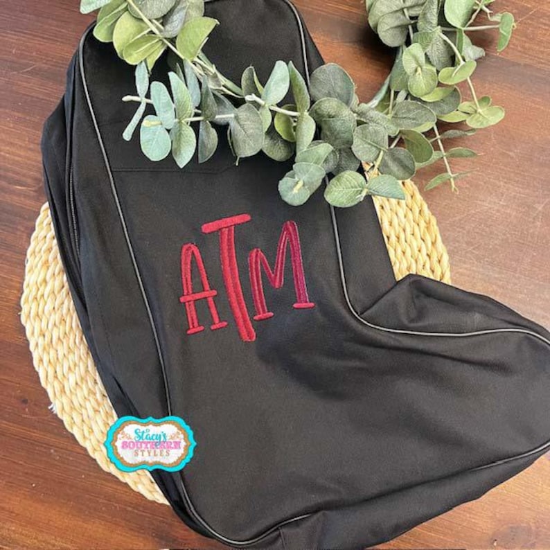 May include: Black bag with a red embroidered "ATM" monogram. The bag has a zipper closure and a light-colored trim. The bag is decorated with a sprig of green leaves and a small, colorful sticker that says "Stacy's Southern Styles".