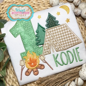 K&ouml;nnte beinhalten: Wei&szlig;es T-Shirt mit Camping-Motiv. Mit einer gr&uuml;nen Zahl 1, einem Zelt, B&auml;umen, einem Lagerfeuer mit Marshmallows und dem Namen "KODIE" in Gr&uuml;n. Enth&auml;lt einen Mond und Sterne.