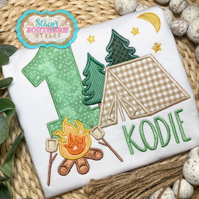 K&ouml;nnte beinhalten: Wei&szlig;es T-Shirt mit Camping-Motiv. Mit einer gr&uuml;nen Zahl 1, einem Zelt, B&auml;umen, einem Lagerfeuer mit Marshmallows und dem Namen "KODIE" in Gr&uuml;n. Enth&auml;lt einen Mond und Sterne.