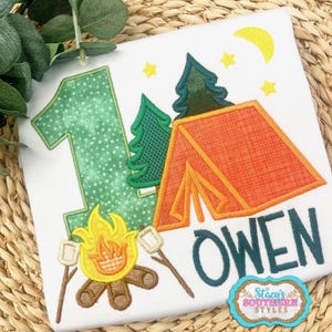 K&ouml;nnte beinhalten: Wei&szlig;es T-Shirt mit Camping-Motiv. Es zeigt die gr&uuml;ne Zahl 1, ein orangefarbenes Zelt, gr&uuml;ne B&auml;ume, ein Lagerfeuer mit Marshmallows und den Namen "OWEN" in T&uuml;rkis. Enth&auml;lt einen gelben Mond und Sterne.