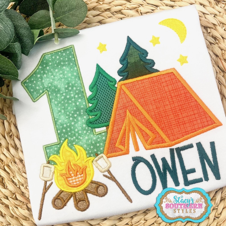 K&ouml;nnte beinhalten: Wei&szlig;es T-Shirt mit Camping-Motiv. Es zeigt die gr&uuml;ne Zahl 1, ein orangefarbenes Zelt, gr&uuml;ne B&auml;ume, ein Lagerfeuer mit Marshmallows und den Namen "OWEN" in T&uuml;rkis. Enth&auml;lt einen gelben Mond und Sterne.