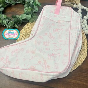 May include: A white and pink boot-shaped bag with a pink handle. The bag features a repeating pattern of cowboy-themed illustrations in pink. The bag is trimmed in pink and has a pocket on the front.
