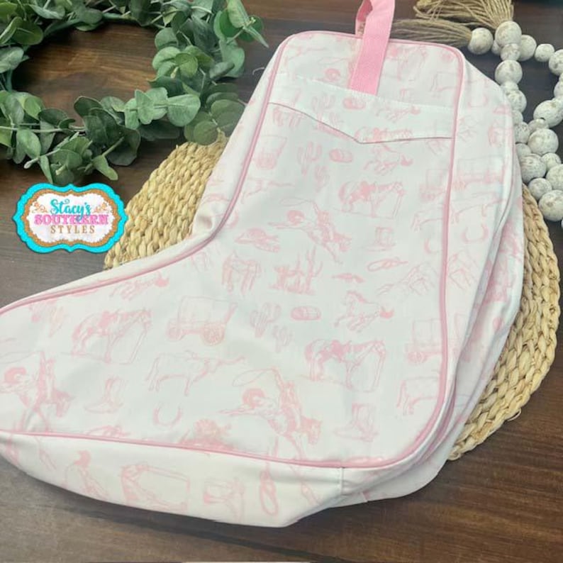 May include: A white and pink boot-shaped bag with a pink handle. The bag features a repeating pattern of cowboy-themed illustrations in pink. The bag is trimmed in pink and has a pocket on the front.