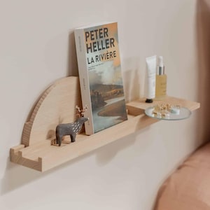 May include: A light-colored wooden floating shelf with a book titled "LA RIVIERE" and a small deer figurine. The shelf also holds a small bottle, a tube of cream, and a small round mirror with decorative items.
