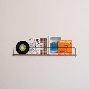 May include: A black vinyl record, a plaid patterned book, a book with a mountain scene, and a book with a man's face and the text "Just a Gigolo" and "Louis Prima" are displayed on a black metal shelf with a wooden base.