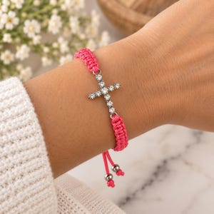 Peut inclure: Un bracelet en cordon tressé rose vif orné d'une croix centrale argentée incrustée de strass transparents étincelants. Le bracelet est doté de cordons de serrage réglables terminés par deux petites perles métalliques argentées.