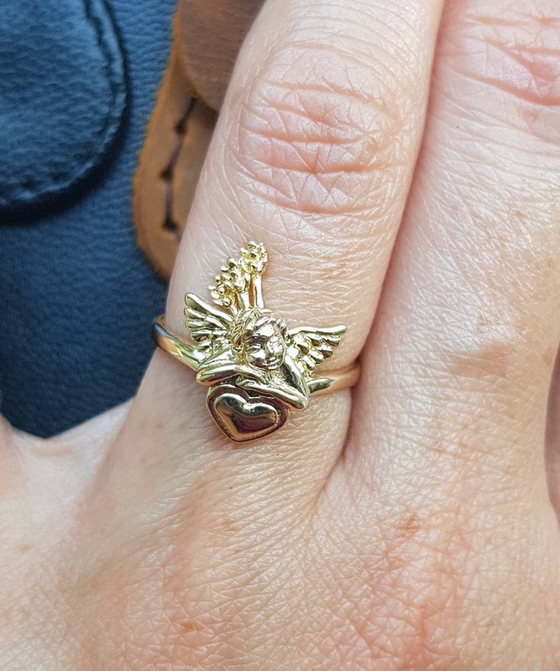 May include: Gold ring featuring a cherub holding a heart. The cherub has wings and a halo. The ring is on a finger.